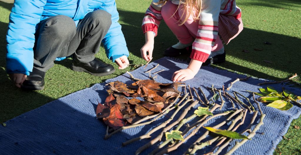 Children making a face by arranging leaves and sticks on a blanket on the grass